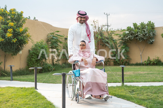Sincerity and kindness between spouses, improving the psychology of people of determination, luxury residential apartments and villas, helping patients with mobility disabilities move easily, a Saudi Gulf Arab man wearing traditional clothing accompanied by his wife in a park, a crippled Saudi woman sitting in a wheelchair