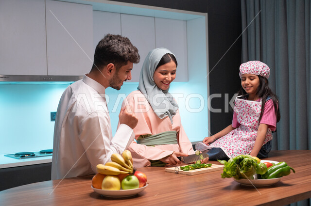 مفهوم النظام الغذائي الصحي، قضاء أوقات ممتعة مع الأبناء، مهارات وفنون الطبخ والتقطيع, ايماءات السعادة والسرور بالتعاون بين افراد العائلة داخل المطبخ, أم عربية خليجية سعودية محجبة تقوم بتعليم ابنتها الطبخ، إعداد وتحضير الطعام بمكونات طبيعية طازجة