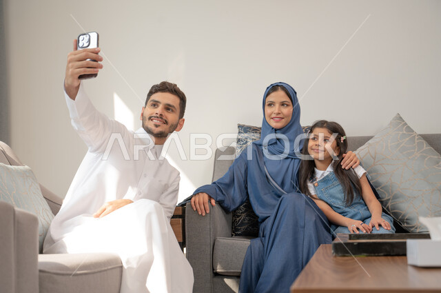 الشعور بالفرح والسرور في عطلة الصيف, أب عربي سعودي خليجي مبتسم يرتدي الثوب التقليدي يلتقط صور السيلفي التذكارية مع أسرته باستخدام الهاتف المحمول، قضاء أوقات عائلية ممتعة في غرفة المعيشة، توثيق اللحظات السعيدة، أجهزة وتقنيات حديثة.