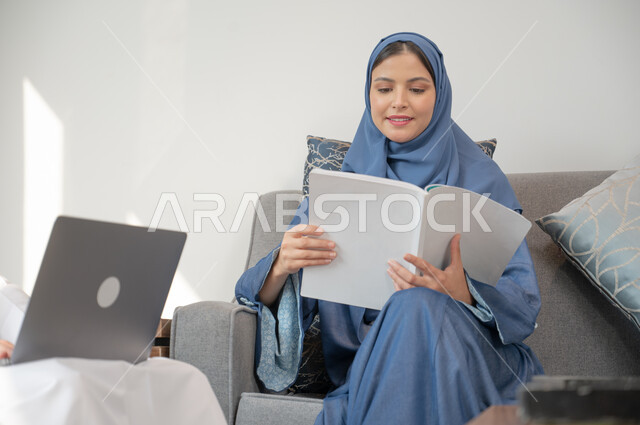  هواية المطالعة والاستكشاف، تنمية وتعزيز المعلومات العامة, إمرأة عربيه سعوديه خليجيه محجبة مبتسمة ترتدي العباءة تجلس على أريكه مريحه في غرفة المعيشة تمسك كتاب, قضاء وقت ممتع في القراءة, الإهتمام بالعلم والثقافه, الحبث والاطلاع على مجالات مختلفة.