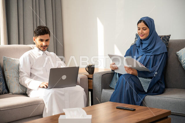 إمرأة تهتم بهواية القراءة ومطالعة الكتب، إجراء مكالمات فيديو عبر الانترنت، رجل عربي خليجي سعودي مبتسم يرتدي الزي التقليدي يمسك جهاز الحاسوب المحمول يجلس على أريكة مريحة مع زوجته، تنمية وتعزيز المعلومات العامة، الضغط والنقر على لوحة المفاتيح بإيماءات التركيز .
