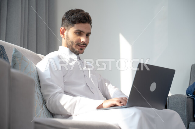 Making video calls over the Internet, a smiling Saudi Arabian Gulf man wearing traditional clothing holding a laptop sitting on a comfortable sofa, e-marketing and working remotely via the laptop, watching movies and series, pressing and clicking on the keyboard with focus gestures