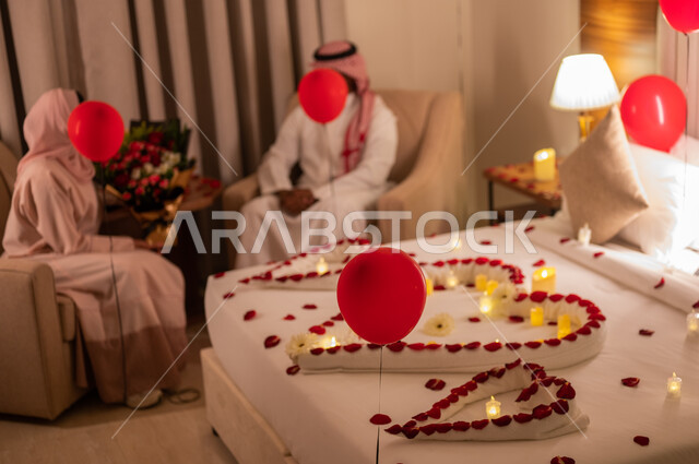 Decorating the room with balloons, a Saudi Gulf Arab couple exchanging conversations and communicating, feelings of love and friendliness, the concept of connection and familiarity, a bed decorated with candles, red roses and balloons to celebrate Valentine’s Day, celebrating the wedding anniversary, paying attention to special occasions