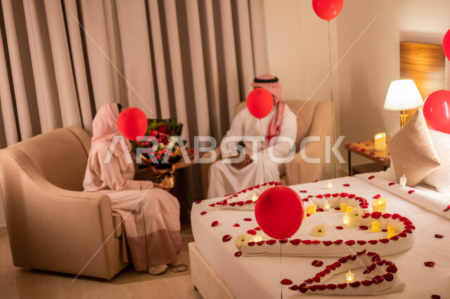 زوجان عربيان خليجيان سعوديان يتبادلان الحديث ويتسامران، مشاعر الحب والود، مفهوم الارتباط والإلفة، سرير مزين بالشموع والورود الحمراء والبالونات للاحتفال بيوم الفلنتاين، الاحتفال بذكرى الزواج، الاهتمام بالمناسبات الخاصة، تزيين غرفة النوم بطريقة رومانسية.