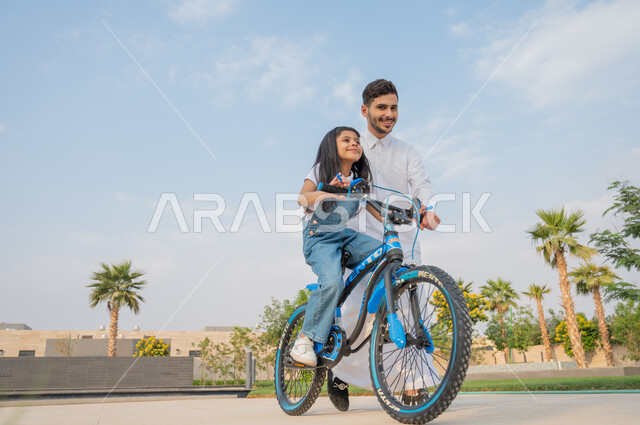 تعليم الأولاد التوازن واللعب في الدراجات الهوائية، رجل عربي خليجي سعودي يرتدي الزي التقليدي يساعد إبنته في قيادة الدراجة في إحدى الحدائق العامة، المشاركة مع الأبناء في الأنشطة الترفيهية، إيماءات الفرح في قضاء أوقات عائلية ممتعة، الطبيعة الخضراء في المملكة.