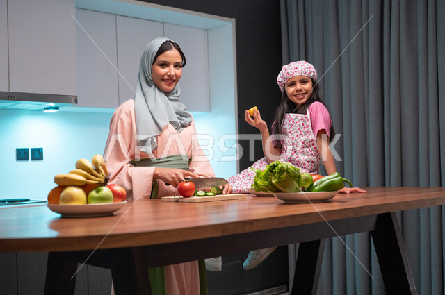النظر الى الكاميرا بايماءات السعادة والسرور، مشاركة الاولاد في إعداد وتحضير الطعام، قضاء أوقات ممتعة مع الأبناء، أم عربية خليجية سعودية محجبة تقوم بتعليم ابنتها فنون الطبخ والطهي داخل المطبخ، مفهوم اتباع النظام الغذائي الصحي