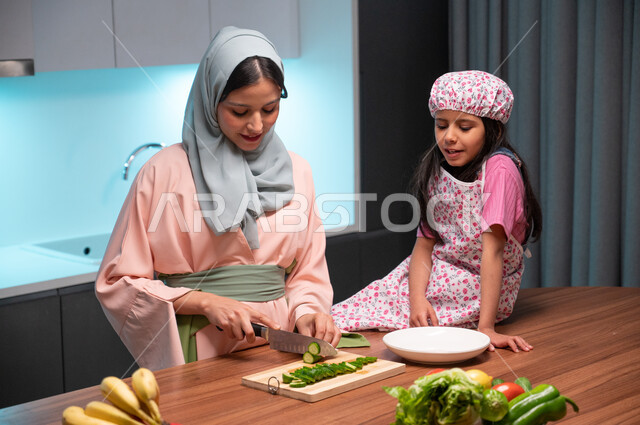 مشاركة الاولاد في إعداد وتحضير الطعام، قضاء أوقات ممتعة مع الأبناء، ايماءات السعادة والسرور بالتعاون بين الفتاة ووالدتها داخل المطبخ, أم عربية خليجية سعودية محجبة تقوم بتعليم ابنتها فنون الطبخ والطهي، مفهوم اتباع النظام الغذائي الصحي