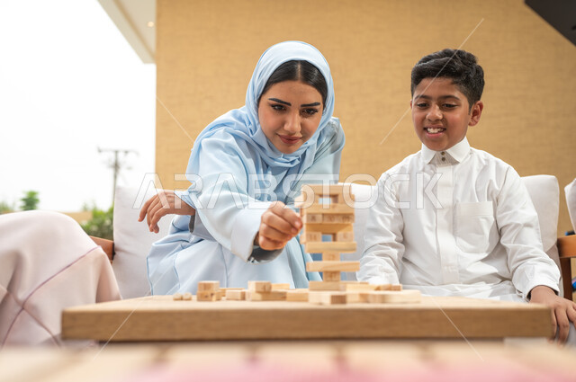 قضاء نزهة ممتعة في اللعب مع الأبناء في الهواء الطلق، صورة مقربة لأم عربية خليجية سعودية مبتسمة تلعب لعبة جينغا بإيماءات الفرح مع ابنها، رص الألواح الخشبية، أنشطة ترفيهية لتنمية مهارات التفكير والذكاء عند الأطفال، ألعاب عائلية مسلية.