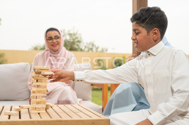 أنشطة ترفيهية لتنمية مهارات التفكير والذكاء عند الأطفال، الاستمتاع في العطلة الصيفية في إحدى المنتجعات، عائلة عربية خليجية سعودية تلعب لعبة جينغا بإيماءات الفرح والمتعة في الهواء الطلق، رص الألواح الخشبية، ألعاب عائلية مسلية.