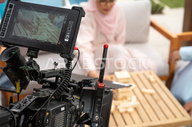 قضاء أوقات ممتعة مع الأبناء في الإجازة الصيفية، ألعاب مسلية لتنمية مهارات التفكير والذكاء عند الأطفال، خلف الكواليس لجلسة تصوير لعائلة عربية خليجية سعودية تلعب لعبة جينغا في الهواء الطلق، رص الألواح الخشبية، لقطة مقربة لكاميرا حديثة ومطورة.