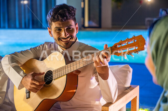 جلسة مسائية طربية في احدى الشاليهات والمنتجعات الراقية، مواهب وفنون موسيقية سعودية، ايماءات السعادة والفرح بممارسة هواية العزف على آلة وترية، صورة مقربة لشاب عربي خليجي سعودي مبتسم يقوم بالعزف على الجيتار، اجواء عائلية سعيدة