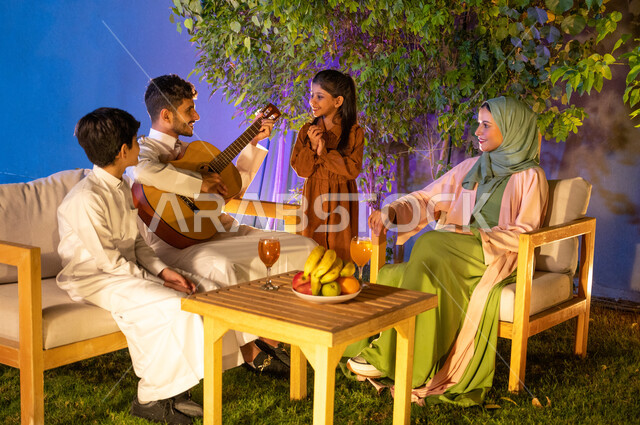 جلسة طربية مسائية في احدى الشاليهات الفاخرة، عائلة عربية خليجية سعودية تقضي وقت ممتع في الهواء الطلق، مفهوم الترابط الاسري، تبادل الاحاديث والدردشة بين الافراد، رجل عربي خليجي سعودي يقوم بالعزف على آلة الجيتارالوترية، اجواء اسرية مليئة بالسعادة والسرور