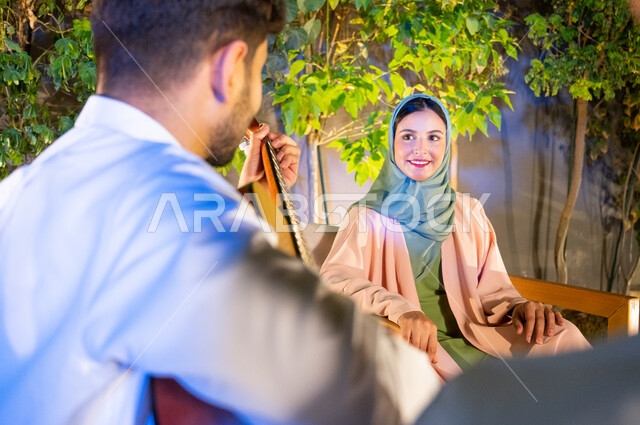 تبادل نظرات الحب بين الزوجين، جلسة مسائية طربية في احدى الشاليهات والمنتجعات الراقية، مواهب وفنون موسيقية سعودية، مفهوم الترابط الاسري وقضاء اوقات عائلية ممتعة، صورة مقربة لشابة عربية خليجية سعودية مبتسمة ترتدي الحجاب بايماءات الاندماج والسرور، اجواء عائلية سعيدة