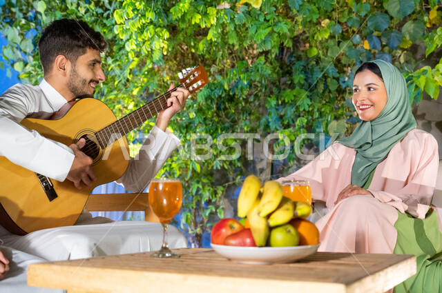 تبادل نظرات الحب بين الزوجين، جلسة مسائية طربية في احدى الشاليهات والمنتجعات الراقية، مواهب وفنون موسيقية سعودية، ايماءات السعادة والفرح بممارسة هواية العزف على آلة وترية، شاب عربي خليجي سعودي مبتسم يقوم بالعزف على الجيتار، اجواء عائلية سعيدة
