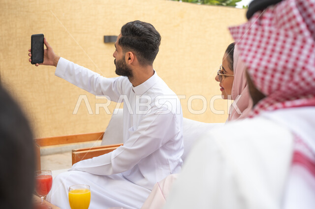 توثيق اللحظات السعيدة بالتقاط الصور التذكارية عبر الهاتف المحمول، صورة مقربة من الخلف لعائلة عربيه خليجيه سعودية تقضي أوقات الفراغ في إحدى الشاليهات، مفهوم الترابط الاسري وصلة الرحم، أجواء مليئة بالحب والود، الاهتمام والاعتناء بالأهل، التنزه في الهواء الطلق