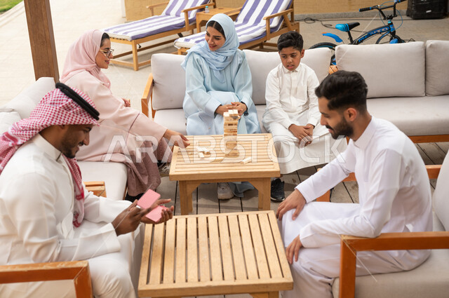قضاء أوقات عائلية ممتعة في أحد الشاليهات في العطلة الصيفية، ممارسة الأنشطة والألعاب الجماعية المسلية لتنمية مهارات التفكير والذكاء، عائلة عربية خليجية سعودية تلعب لعبة جينغا في الهواء الطلق، رجلان سعوديان يلعبان لعبة الورق بإيماءات التركيز والتحدي.