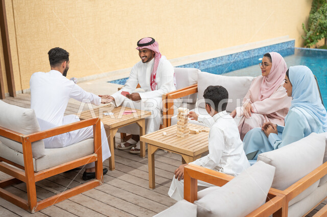 ممارسة الأنشطة والألعاب المسلية لتنمية مهارات التفكير والذكاء، قضاء أوقات عائلية ممتعة في الإجازة الصيفية في إحدى المنتجعات، عائلة عربية خليجية سعودية تلعب لعبة جينغا في الهواء الطلق، رجلان سعوديان يلعبان لعبة الورق بإيماءات السعادة، مفهوم التركيز والتحدي.