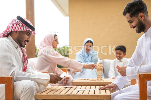 ممارسة الأنشطة والألعاب المسلية لتنمية مهارات التفكير والذكاء، قضاء أوقات عائلية ممتعة في الإجازة الصيفية في إحدى المنتجعات، عائلة عربية خليجية سعودية تلعب لعبة جينغا في الهواء الطلق، رجلان سعوديان يلعبان لعبة الورق بإيماءات السعادة، مفهوم التركيز والتحدي.