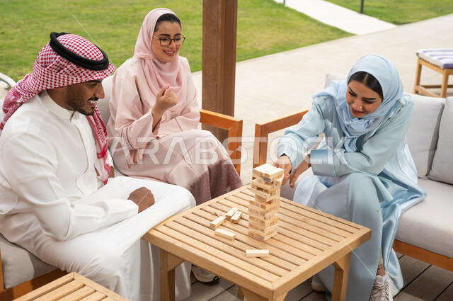 إيماءات المتعة والسعادة في أحد الشاليهات في العطلة الصيفية، ممارسة الأنشطة والألعاب الجماعية المسلية لتنمية مهارات التفكير والذكاء، عائلة عربية خليجية سعودية تلعب لعبة جينغا في الهواء الطلق، رص القطع والألواح الخشبية