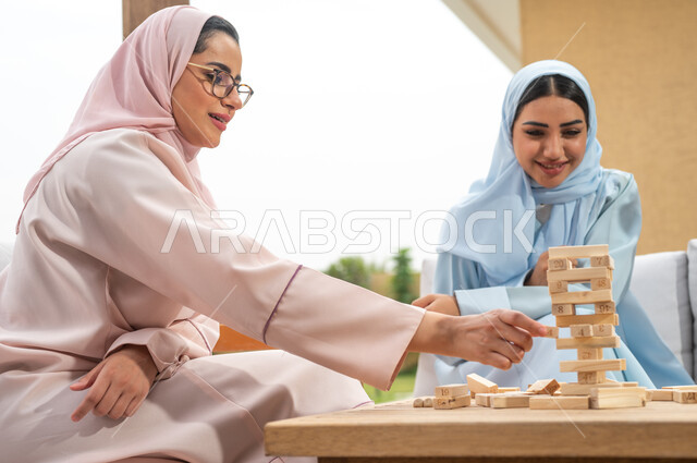 ممارسة الأنشطة والألعاب المسلية مع الأصدقاء، إيماءات المتعة والسعادة في أحد الشاليهات في العطلة الصيفية، صورة مقربة لسيدتان عربيتان خليجيتان سعوديتان تلعبان لعبة جينغا في الهواء الطلق، رص القطع والألواح الخشبية