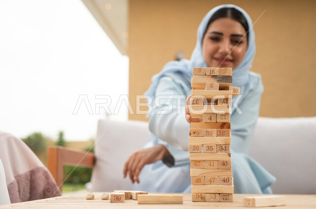 رص القطع والألواح الخشبية، ممارسة الأنشطة والألعاب المسلية في المنتجع، المتعة والسعادة في أحد الشاليهات في العطلة الصيفية، صورة مقربة لسيدة عربية خليجية سعودية تلعب لعبة جينغا في الهواء الطلق بإيماءات الاستمتاع