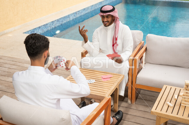 مفهوم التركيز والتحدي في لعب الشدة، قضاء وقت ممتع في ممارسة الأنشطة والألعاب المسلية، الاستمتاع والتسلية في أحد الشاليهات في العطلة الصيفية، صورة مقربة لرجلين عربيين خليجيين سعوديين يلعبان لعبة الورق بإيماءات السعادة والسرور 