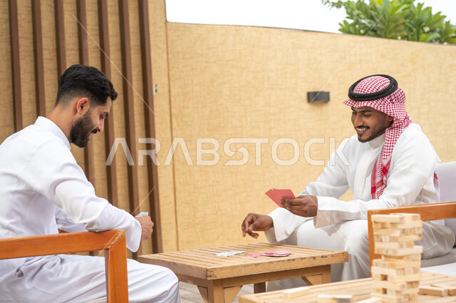 مفهوم التركيز والتحدي في لعب الشدة، قضاء وقت ممتع في ممارسة الأنشطة والألعاب المسلية، الاستمتاع والتسلية في أحد الشاليهات في العطلة الصيفية، صورة مقربة لرجلين عربيين خليجيين سعوديين يلعبان لعبة الورق بإيماءات السعادة والسرور 