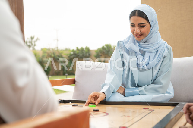 أجواء صيفية سعيدة، قضاء أوقات ممتعة في الهواء الطلق، التسلية والترفيه في إحدى الشاليهات السياحية، مفهوم التحدي والتركيز، ألعاب شعبية في المملكة، صورة مقربة لامرأة عربية خليجية سعودية محجبة تلعب بلعبة الكيرم (البليارد) المسلية في الخارج.