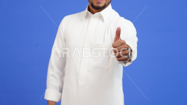 إشارات حركات الاعجاب والقبول والموافقة, بورتريه مقرب لرجل عربي خليجي سعودي يرتدي الثوب التقليدي يرفع اصبع الابهام للأعلى بإيماءات علامة الجودة والرضا، خلفية زرقاء