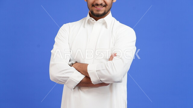 الوقوف بثقة والاهتمام بالمظهر الخارجي, بورتريه مقرب لرجل عربي خليجي سعودي مبتسم يرتدي الثوب التقليدي يقف بأيدي متقاطعة بإيماءات وتعابير السرور والسعادة، خلفية زرقاء