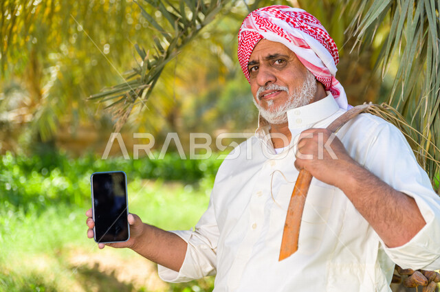 مزارع سعودي خليجي يحمل بيده الهاتف المحمول ، شراء التمر اونلاين ، الشراء عن طريق الانترنت ، البيع اونلاين ، الموسم الزراعي ، موسم جني محصول التمور في المناطق الزراعية ، مزارع النخيل المثمرة في المملكة العربية السعودية ، مزارع لانتاج التمور ، المناطق الزرا