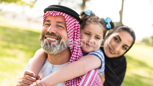 التنزه في الهواء الطلق في وضح النهار، مفهوم الترابط الأسري، قضاء اوقات عائلية ممتعة، صورة مقربة لرجل عربي خليجي سعودي يرتدي الشماغ والثوب التقليدي يحمل ابنته على ظهره بجانب زوجته في احدى الحدائق العامة، ايماءات وتعابير السعادة والسرور لاجتماع العائلة