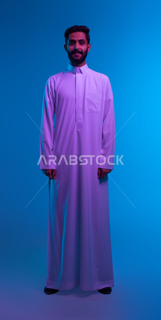 الاهتمام بالمظهر الخارجي، الوقوف باستقامة وثبات، بورتريه مقرب لشاب عربي سعودي خليجي مبستم يرتدي الثوب التقليدي ينظر الى الكاميرا بايماءات الثقة بالنفس و البشاشة، صورة شخصية للوثائق الرسمية, مفهوم الرجولة والأناقه، صورة الجسم بالطول الكامل، خلفية زرقاء
