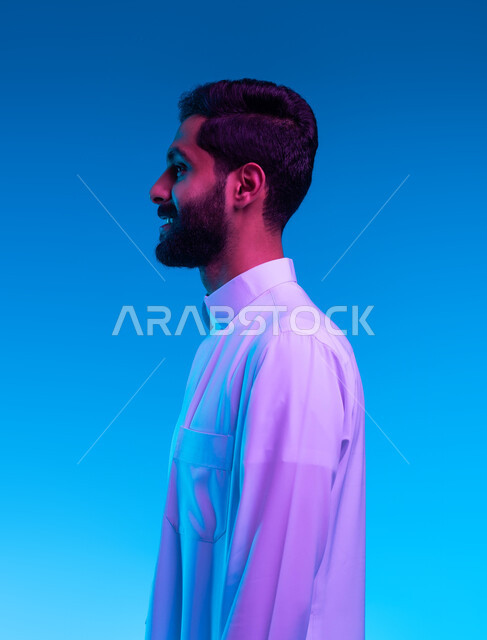 التزين و التجمل و الاهتمام بالمظهر الخارجي و الأناقه، مفهوم الرجولة و الانضباط و الثبات، بورتريه مقرب من الجانب لشاب عربي سعودي خليجي مبستم يرتدي الثوب التقليدي يقف بايماءات السرور و الثقة بالنفس و البشاشة، خلفية زرقاء