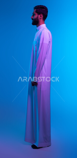 التزيين و التجمل و الاهتمام بالمظر الخارجي، بورتريه من الجانب لشاب عربي سعودي خليجي يرتدي الثوب التقليدي يضع يديه على جانبه بإيماءات الاعتزاز بالنفس و الانضباط، الوقوف باستقامه و ثقة، صورة الجسم بالطول الكامل، خلفية زرقاء