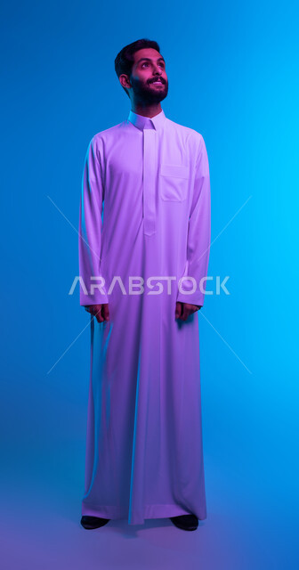 Adornment and attention to the external appearance, a portrait of a smiling young Saudi Arabian Gulf man wearing the traditional Saudi dress, placing his hands on his side, looking upward, standing with gestures of pleasure, discipline, and self-confidence.   Full length body portrait, blue background
