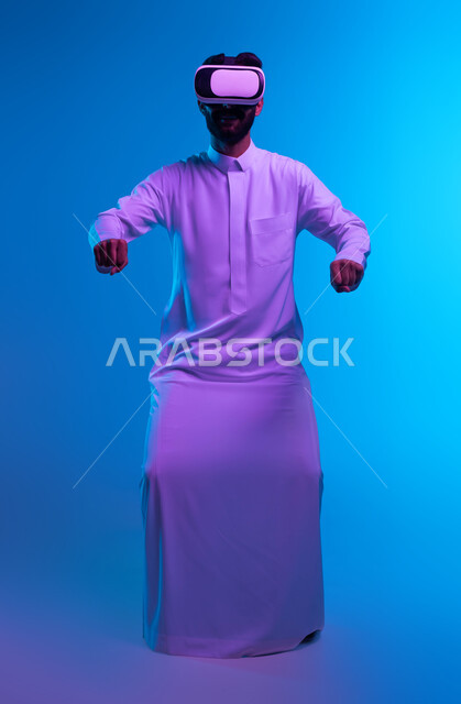 محاكاة ركوب سيارة ثري دي في العالم الخيالي، بورتريه لشاب عربي خليجي سعودي يرتدي الثوب التقليدي ونظارة الواقع الإفتراضي ثلاثية الأبعاد يقوم باللعب في ألعاب الفيديو، تقنيات وأجهزة تكنولوجية، إيماءات التفاعل بألعاب قيادة الموتورسايكل، صورة الجسم بالكامل، خلفية زرقاء