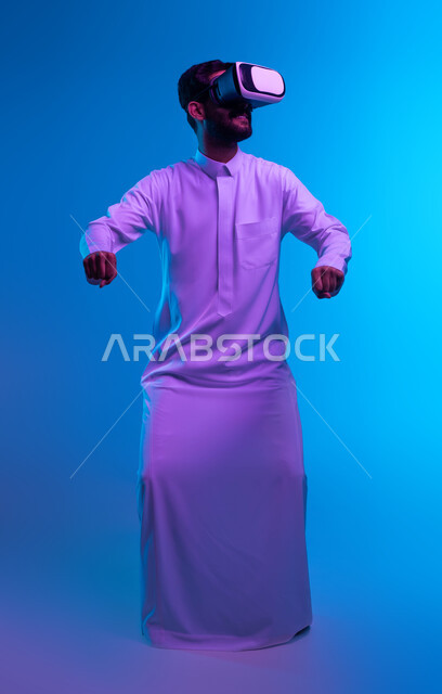 إيماءات يدين تدل على قيادة السيارة، أجهزة وتقنيات تكنولوجية، محاكاة ركوب سيارات ثري دي في العالم الخيالي، بورتريه لشاب عربي خليجي سعودي يرتدي الثوب التقليدي ونظارة الواقع الإفتراضي ثلاثية الأبعاد يقوم باللعب في ألعاب الفيديو، صورة الجسم بالكامل، خلفية زرقاء