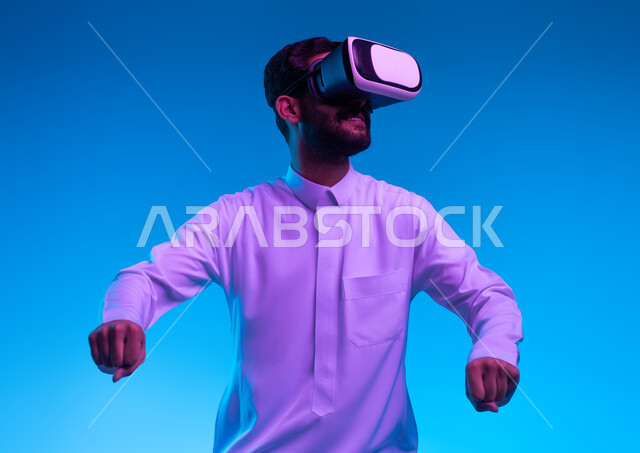 محاكاة ركوب موتورسايكل ثري دي في العالم الخيالي، بورتريه مقرب لشاب عربي خليجي سعودي يرتدي الثوب التقليدي ونظارة الواقع الإفتراضي ثلاثية الأبعاد يقوم باللعب في ألعاب الفيديو، تقنيات وأجهزة تكنولوجية، إيماءات يدين تدل على السعادة بألعاب قيادة الدراجات، خلفية زرقاء