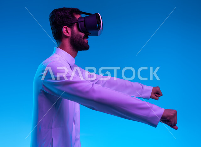محاكاة ركوب موتورسايكل ثري دي في العالم الخيالي، بورتريه مقرب من الجانب لشاب عربي خليجي سعودي يرتدي الثوب التقليدي ونظارة الواقع الإفتراضي ثلاثية الأبعاد يقوم باللعب في ألعاب الفيديو، تقنيات وأجهزة تكنولوجية، إيماءات السعادة بألعاب قيادة الدراجات، خلفية زرقاء