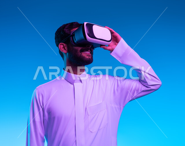 التطلع الى المستقبل و محاكاة العالم الخيالي، بورتريه مقرب لشاب عربي خليجي سعودي مبتسم يرتدي الزي التقليدي و نظارة الواقع الإفتراضي ثلاثية الأبعاد الفريدة ويمسكها بيده، ايماءات التفاعل والتسلية بألعاب الثري دي، تقنيات و أجهزة الكترونية حديثة، خلفية زرقاء