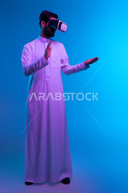 تقنيات حديثة الكترونية وأجهزة تكنولوجية، التفاعل ومحاكاة العالم الخيالي, بورتريه لشاب عربي خليجي سعودي يرتدي الثوب التقليدي و نظارات الواقع الافتراضي بايماءات الاندماج ومحاولة امساك ولمس شيء ما, صورة الجسم بالطول الكامل، خلفية زرقاء