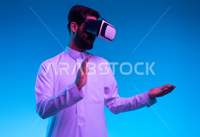 التفاعل ومحاكاة العالم الخيالي, بورتريه مقرب لشاب عربي خليجي سعودي يرتدي الثوب التقليدي و نظارات الواقع الافتراضي بايماءات الاندماج ومحاولة امساك ولمس شيء ما, تقنيات حديثة الكترونية وأجهزة تكنولوجية، خلفية زرقاء