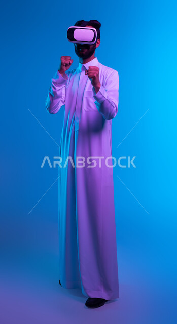 ممارسة رياضة الدفاع عن النفس بتقنيات تكنولوجية، التدرب على الحركات القتالية عبر ألعاب الفيديو، بورتريه لشاب عربي خليجي سعودي يرتدي الثوب التقليدي ونظارة الواقع الافتراضي بإيماءات الملاكمة والمصارعة، تفريغ الطاقة السلبية، صورة الجسم بالكامل، خلفية زرقاء