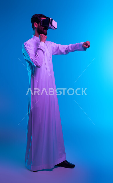صورة الجسم بالكامل، تفريغ الطاقة السلبية، ممارسة رياضة الدفاع عن النفس بتقنيات تكنولوجية، التدرب على الحركات القتالية عبر ألعاب الفيديو، بورتريه جانبي لشاب عربي خليجي سعودي يرتدي الثوب التقليدي ونظارة الواقع الافتراضي بإيماءات الملاكمة والمصارعة، خلفية زرقاء