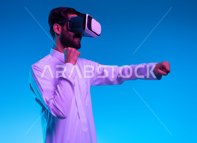 التدرب على الحركات القتالية من خلال ألعاب الفيديو، تفريغ الطاقة السلبية، ممارسة رياضة الدفاع عن النفس بتقنيات تكنولوجية، بورتريه جانبي مقرب لشاب عربي خليجي سعودي يرتدي الثوب التقليدي ونظارات الواقع الافتراضي بإيماءات الملاكمة والمصارعة، خلفية زرقاء