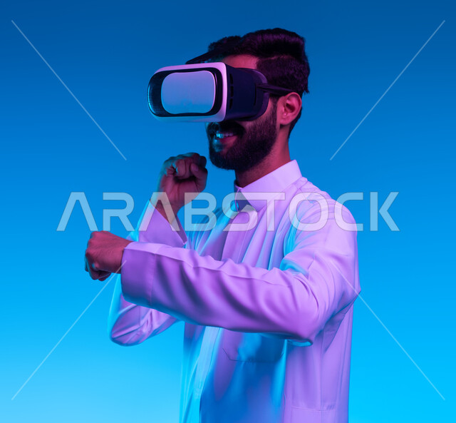 التعلم على الحركات القتالية من خلال ألعاب الفيديو، تفريغ الطاقة السلبية، ممارسة رياضة الدفاع عن النفس بتقنيات تكنولوجية، بورتريه جانبي مقرب لشاب عربي خليجي سعودي يرتدي الثوب التقليدي ونظارات الواقع الافتراضي بإيماءات الملاكمة والمصارعة، خلفية زرقاء