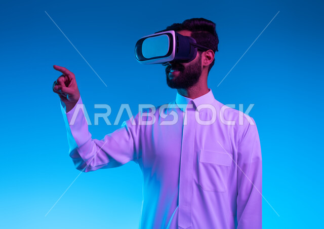 ايماءات الضغط واللمس، بورتريه مقرب لرجل عربي خليجي سعودي يرتدي الثوب التقليدي ونظارات في آر ثلاثية الأبعاد يشير بأصبع السبابة الى شئ ما يشاهده بالواقع الإفتراضي، محاكاة العالم الخيالي، اشارات النقر على شاشه هولوجرامية، خلفية زرقاء