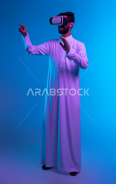 Simulating the imaginary world, touch and pressure movements, a portrait of a Saudi Gulf Arab man wearing a traditional thobe and 3D VR glasses, standing straight and pointing with his index finger at something he is watching in virtual reality, click signals on a holographic screen, full body image, blue background