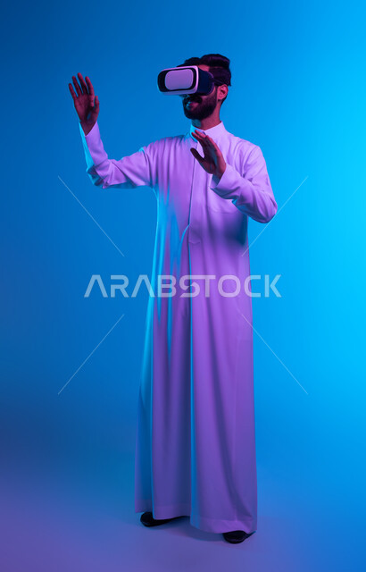Merging and trying to hold and touch something, interacting and simulating the imaginary world, portrait of a Saudi Gulf Arab young man wearing a traditional dress and virtual reality glasses with self-defense gestures, modern techniques and technological devices, full-length body image, blue background